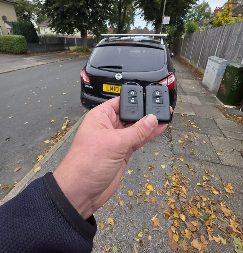 Hand holding two new remote keys in front of a Nissan Qashqai on a leafy Coventry street
